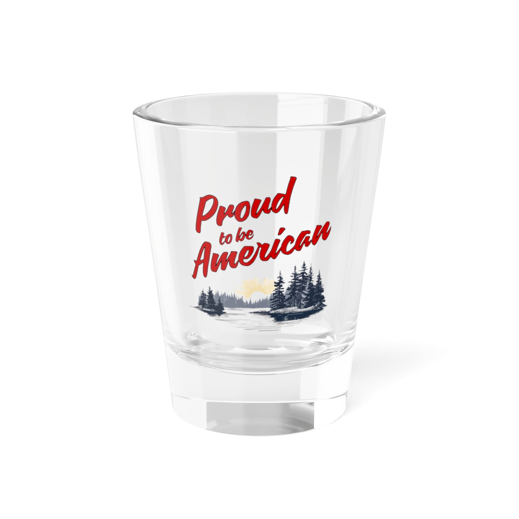A clear glass shot glass with 'Proud to be American' printed in red and a small depiction of an American flag and landscape in the background.
